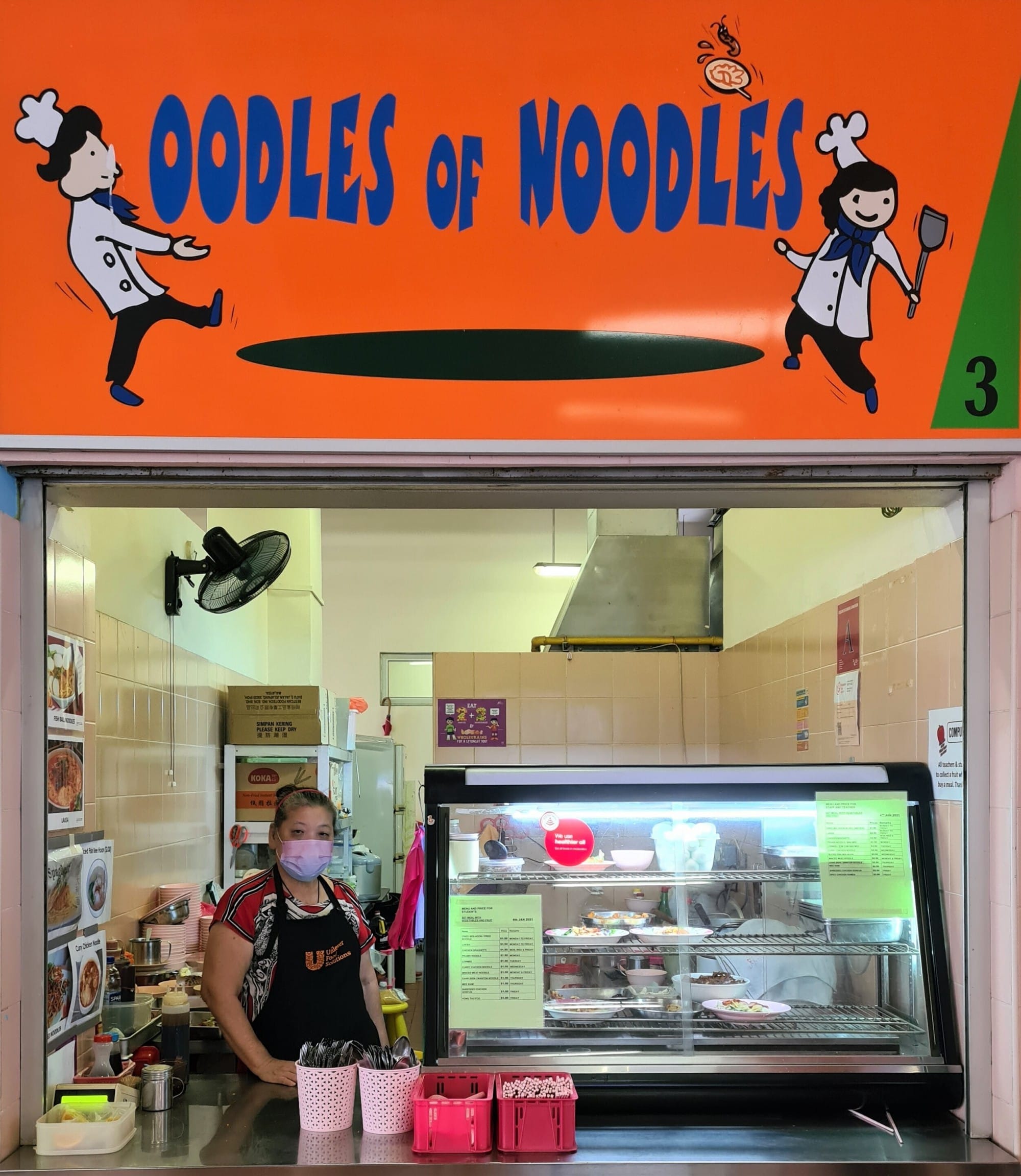 A food stall called Oodles of Noodles with a masked worker standing behind a display counter of dishes.